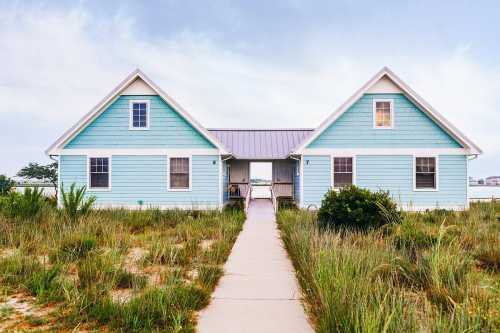 A charming blue beach house with a pathway surrounded by grass, leading to a serene coastal view.