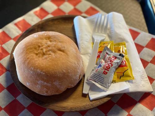 A wooden plate with a soft bread roll, condiments, and a fork on a checkered tablecloth.