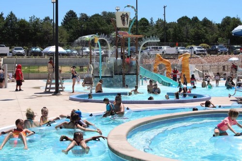 A busy water park scene with children playing in pools and on slides under a clear blue sky.