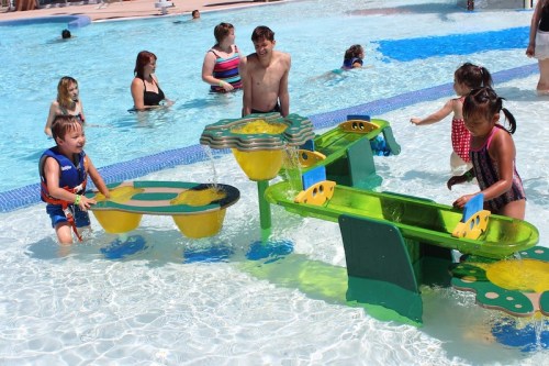 Children play in a water park, interacting with colorful water features while adults supervise nearby.