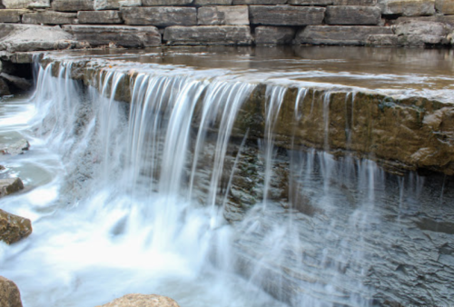 A serene waterfall cascading over rocky ledges into a calm pool below, surrounded by natural stone formations.