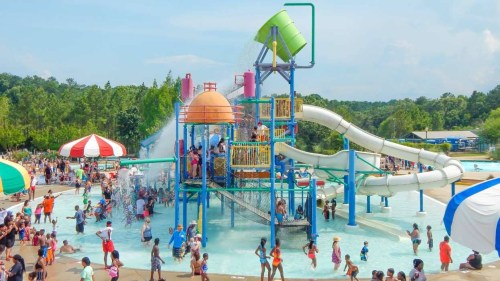 A busy water park with colorful slides, a splash pad, and many people enjoying the water on a sunny day.