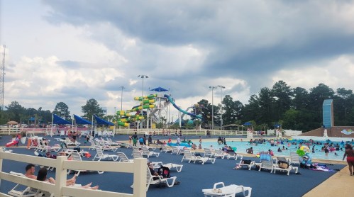 A busy water park with a large pool, water slides, and many people enjoying the sunny day under cloudy skies.