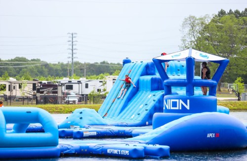 A blue inflatable water park with slides and climbing structures, set on a lake with trees and campers in the background.