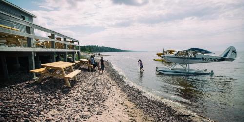 A seaplane is parked on the water near a beach, with people walking along the shore and a wooden deck in the background.