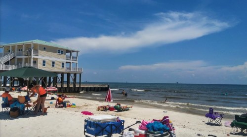 A sunny beach scene with a pier, colorful beach chairs, and people enjoying the water under a blue sky.