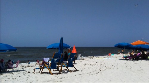 A sunny beach scene with blue umbrellas, lounge chairs, and people enjoying the shoreline and ocean waves.