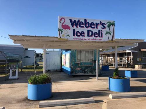 A colorful ice deli stand with a sign reading "Weber's Ice Deli," surrounded by planters and a clear blue sky.