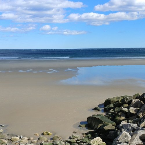 A serene beach scene with gentle waves, a clear blue sky, and scattered clouds reflecting on the water.