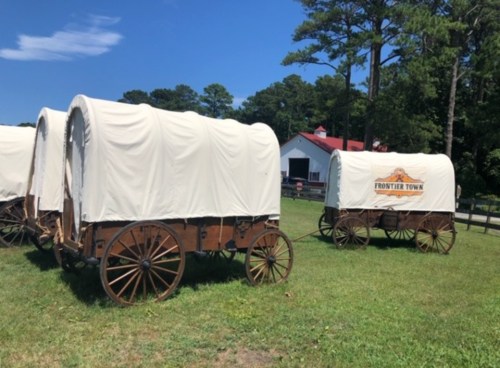 Covered wagons parked on green grass, with a barn and trees in the background under a clear blue sky.
