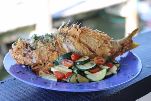 A whole fried fish garnished with herbs, served on a plate with a colorful vegetable medley of zucchini and cherry tomatoes.
