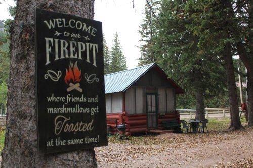 A sign reading "Welcome to our Firepit" next to a cozy cabin surrounded by trees, inviting friends to gather.