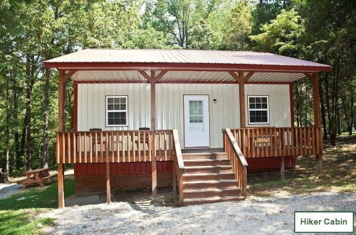 A small cabin with a red roof and wooden porch, surrounded by trees in a natural setting.