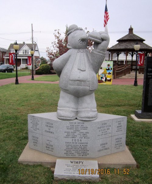 A stone statue of Wimpy from Popeye, holding a hamburger, stands on a pedestal with engraved names and dates.