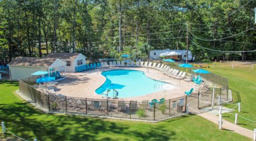 Aerial view of a pool surrounded by lounge chairs and trees, with a small building nearby and a fenced area.
