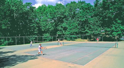 Two children playing tennis on an outdoor court surrounded by trees under a clear blue sky.