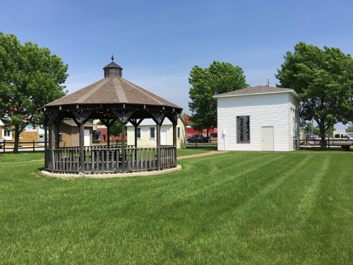 A wooden gazebo in a grassy area, with a white building and trees in the background under a clear blue sky.
