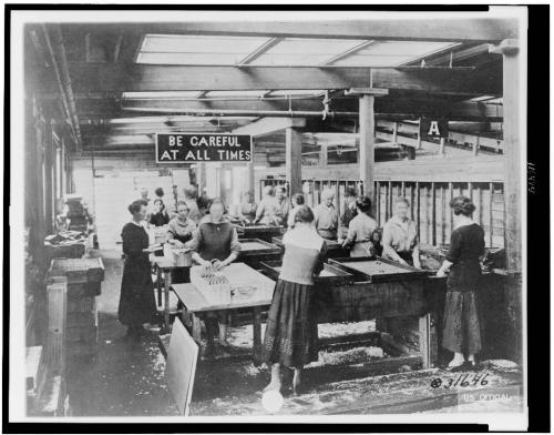 Women working in a factory setting, assembling products at long tables, with a sign reading "Be Careful at All Times."