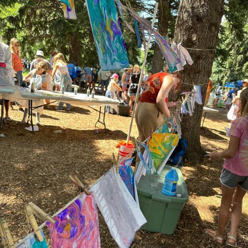 A vibrant outdoor art event with people painting and colorful artworks hanging on a line between trees.