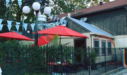 A cozy outdoor dining area with red umbrellas, surrounded by greenery and festive bunting, next to a wooden building.