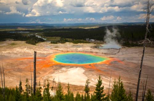 Aerial view of Yellowstone's Grand Prismatic Spring, showcasing vibrant colors and surrounding landscape under a cloudy sky.