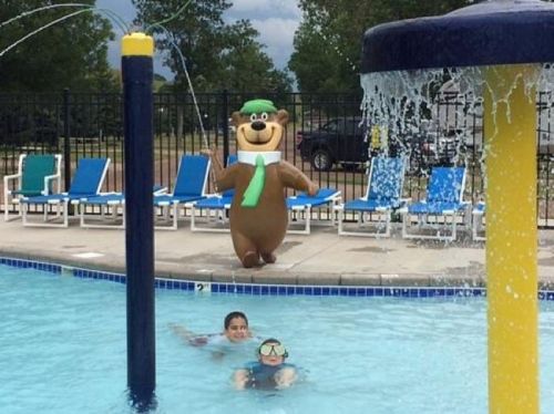 A bear mascot in a green hat stands by a pool while two children swim nearby.