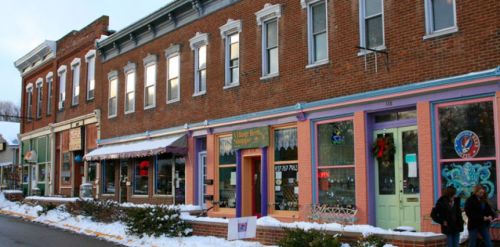 Charming brick storefronts with colorful accents, snow-covered ground, and people walking in a quaint town setting.