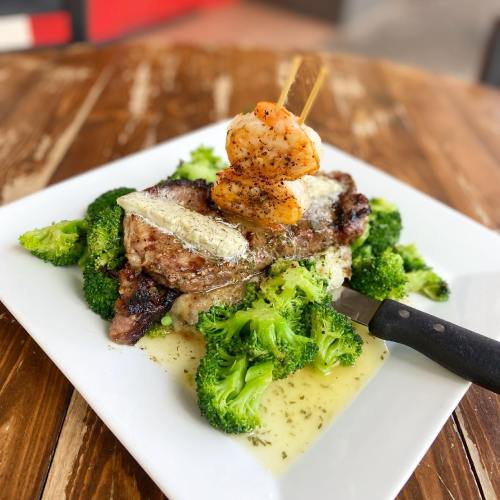 A plate of grilled steak topped with shrimp, served on a bed of broccoli with a drizzle of sauce.