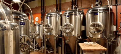A row of shiny stainless steel fermentation tanks in a brewery, with wooden tables and brick walls in the background.