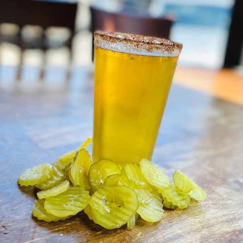 A glass of yellow drink with a rim of seasoning, surrounded by slices of pickles on a wooden table.