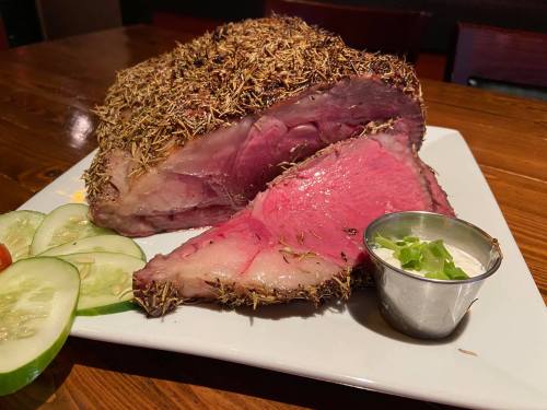 A beautifully cooked roast beef with a herb crust, served with sliced cucumbers and a small cup of sauce.