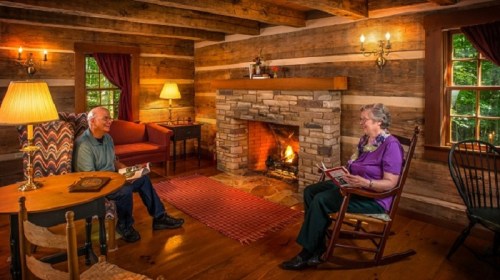 A cozy log cabin interior with two people reading by a fireplace, warm lighting, and rustic wooden decor.