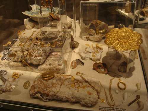 Display of ancient jewelry and artifacts on stone, including gold pieces and ceramic items in a museum setting.