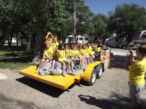 A group of children in yellow shirts sitting on a trailer, smiling and posing for a photo in a park setting.