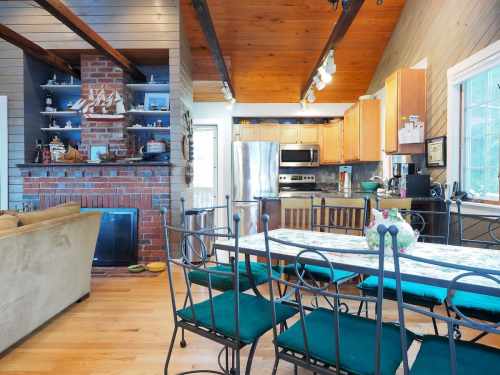 Cozy dining area with a wooden table, green chairs, and a view of a modern kitchen and brick fireplace.