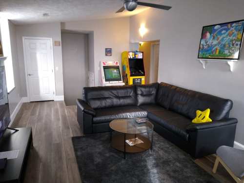 A modern living room with a black sectional sofa, glass coffee table, and arcade games in the background.