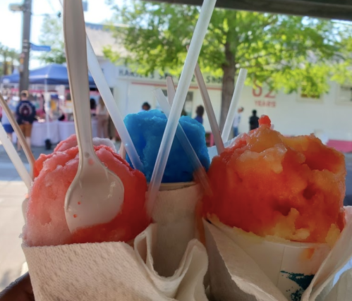 Three colorful snow cones in cups, featuring red, blue, and orange flavors, held with spoons against a sunny backdrop.