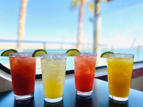 Four colorful cocktails with lime slices, set against a sunny waterfront view.