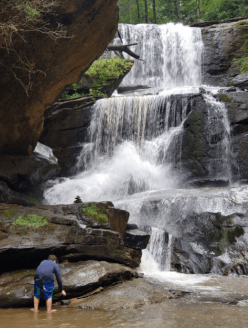 A person stands in shallow water near a cascading waterfall surrounded by rocky terrain and lush greenery.