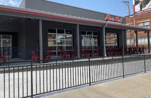 Exterior of a restaurant with a modern design, featuring red chairs and a fenced patio area on a sunny day.