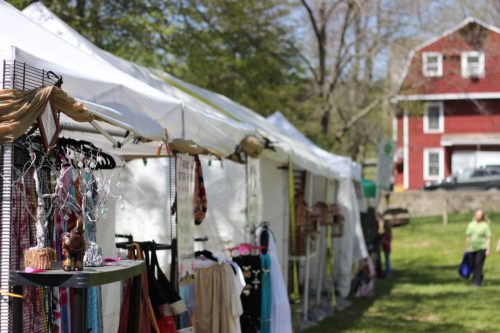 A row of white tents displaying clothing and accessories at an outdoor market, with a red house in the background.