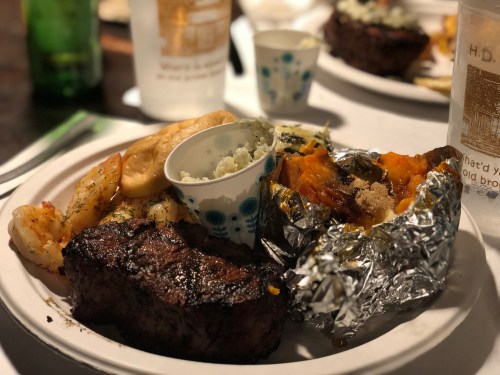 A plate of food featuring grilled steak, shrimp, a baked potato, and a small cup of coleslaw.