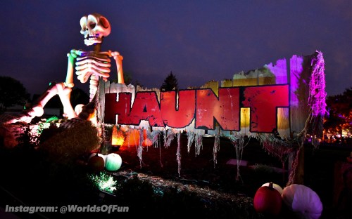 A large skeleton beside a colorful "HAUNT" sign, illuminated with purple and green lights in a spooky setting.