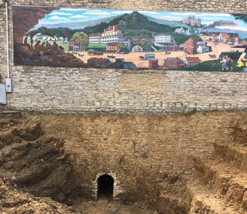 A mural depicting a town scene above a large excavation site with a tunnel entrance at the bottom.