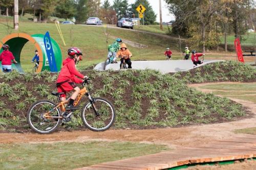 Children riding bikes on a dirt track with hills and colorful banners in a park setting.