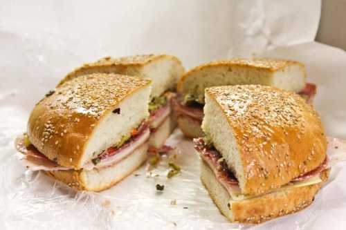 Four sesame seed sandwich rolls filled with layers of meats and condiments, partially cut and arranged on white paper.