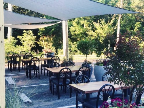 Outdoor dining area with empty tables under shade sails, surrounded by greenery and potted plants.