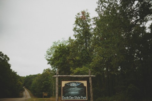 Sign for Devils Den State Park surrounded by trees, with a road visible in the background under a cloudy sky.