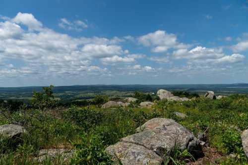 A scenic view of rolling hills and a blue sky dotted with clouds, featuring large rocks in the foreground.