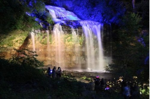 A illuminated waterfall cascades down rocks, with people gathered nearby, surrounded by lush greenery at night.